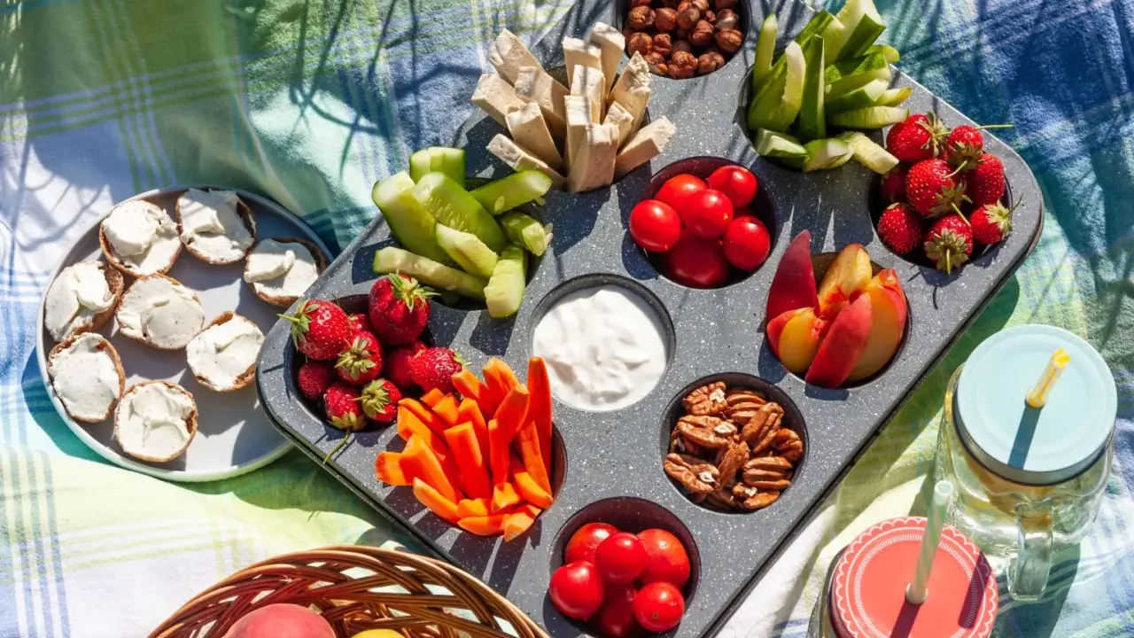 Leckere Picknick-Snacks: Erdbeeren, Gurken, Karotten, Nüsse, Tomaten, Pfirsiche und Brotscheiben mit Frischkäse auf einer Decke.