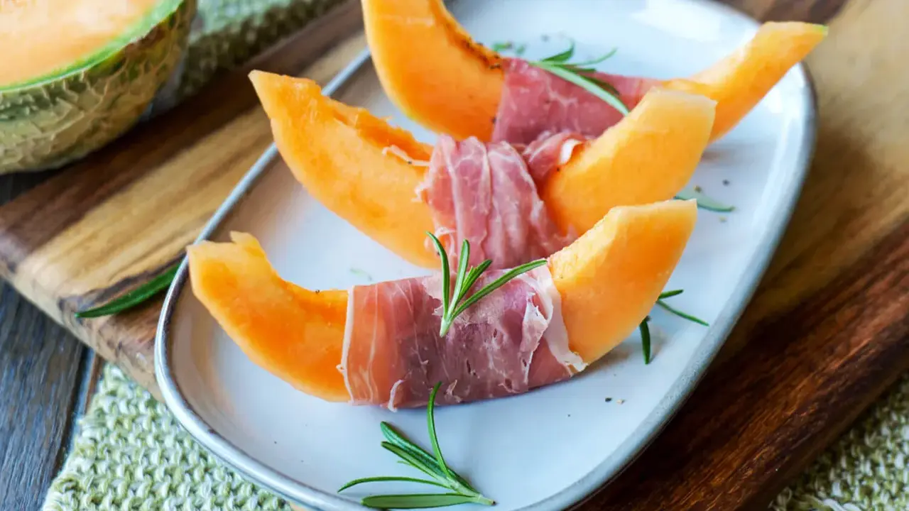 Melone mit Schinken, garniert mit Rosmarin, auf einem Teller angerichtet. Ein halber Melone liegt daneben auf einem Holzbrett.