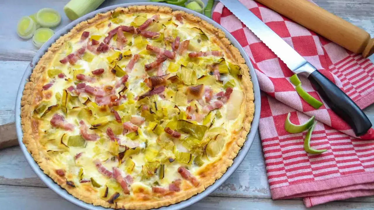Herzhafte Quiche mit Lauch und Speck, perfekt für herzhafte Quiche Rezepte. Daneben liegen Lauch, ein Messer und ein Nudelholz.