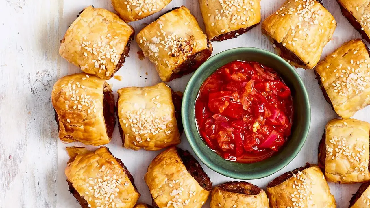 Goldbraune Würstchenschnecken mit Sesam, daneben eine Schale mit roter Chutney-Sauce. Perfekt, um schnelles Fingerfood am Vortag zuzubereiten.