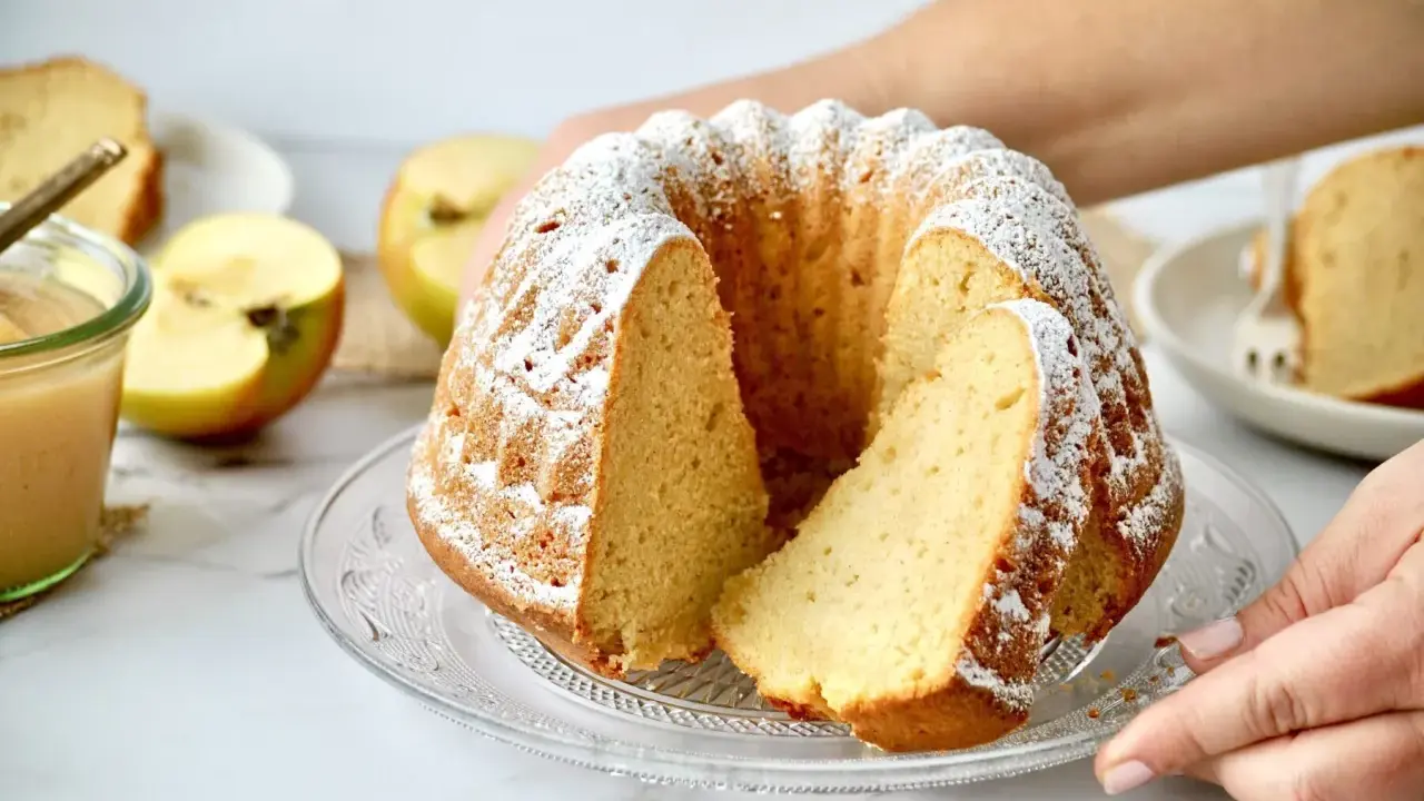 Ein saftiger Kuchen mit Apfelmus, bestäubt mit Puderzucker. Daneben liegen Äpfel und ein Glas Apfelmus.