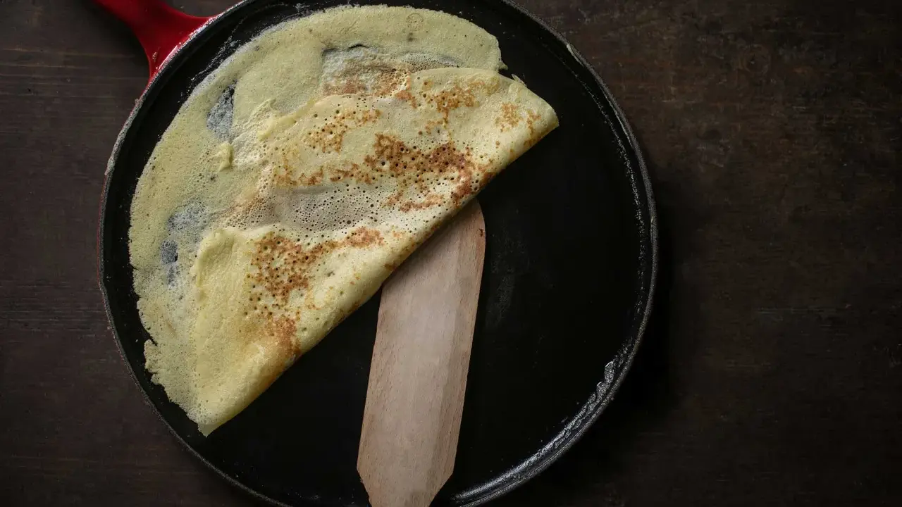 Ein goldbrauner Crêpe wird in einer Pfanne mit rotem Griff gefaltet. Das **Crêpes Teig Rezept Original** gelingt so perfekt.