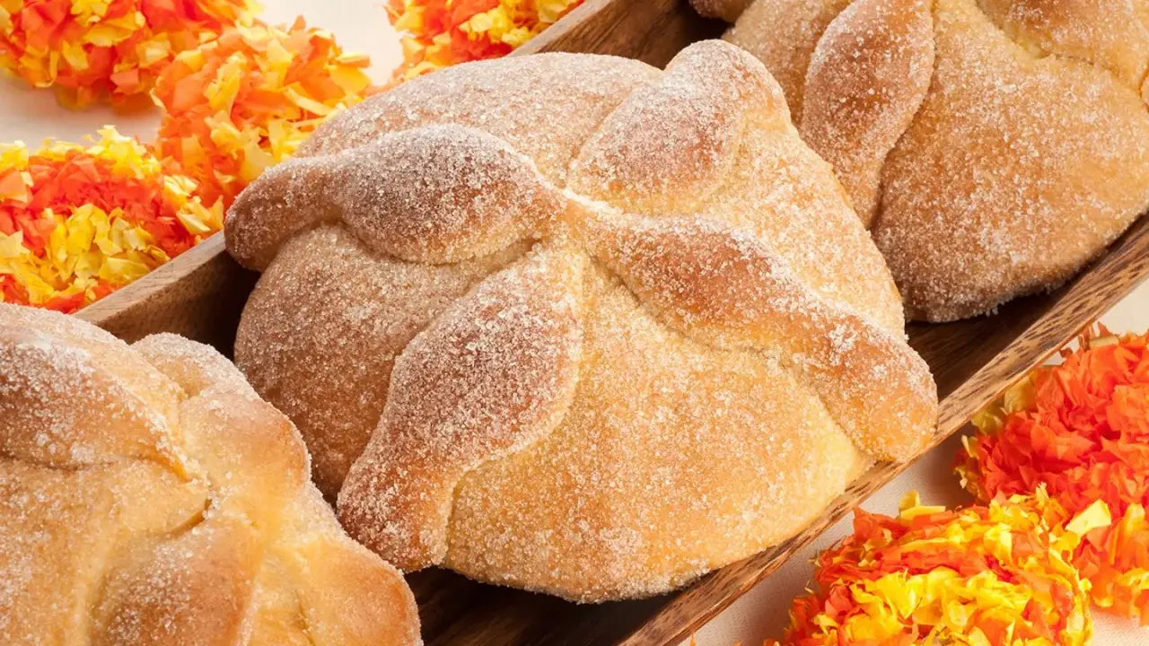 Pan de muerto, süßes Brot mit Zucker bestreut, liegt in einer Holzschale, umgeben von bunten Papierblumen.