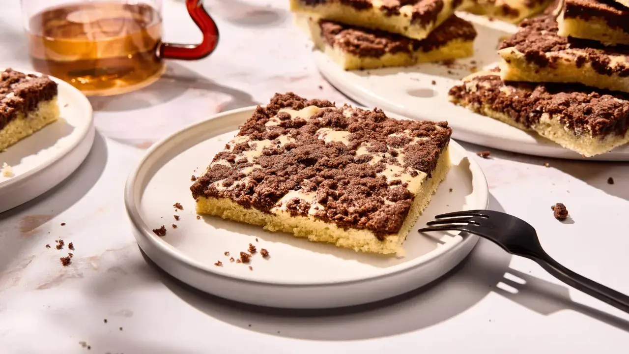 Ein Stück versunkener Streuselkuchen mit Schokostreuseln auf einem Teller, daneben eine Gabel und ein Glas Tee.