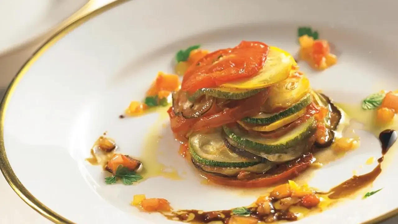 Ein kunstvoll geschichtetes Ratatouille-Rezept, inspiriert vom Film, mit Tomaten, Zucchini und Aubergine auf einem Teller mit goldenem Rand.