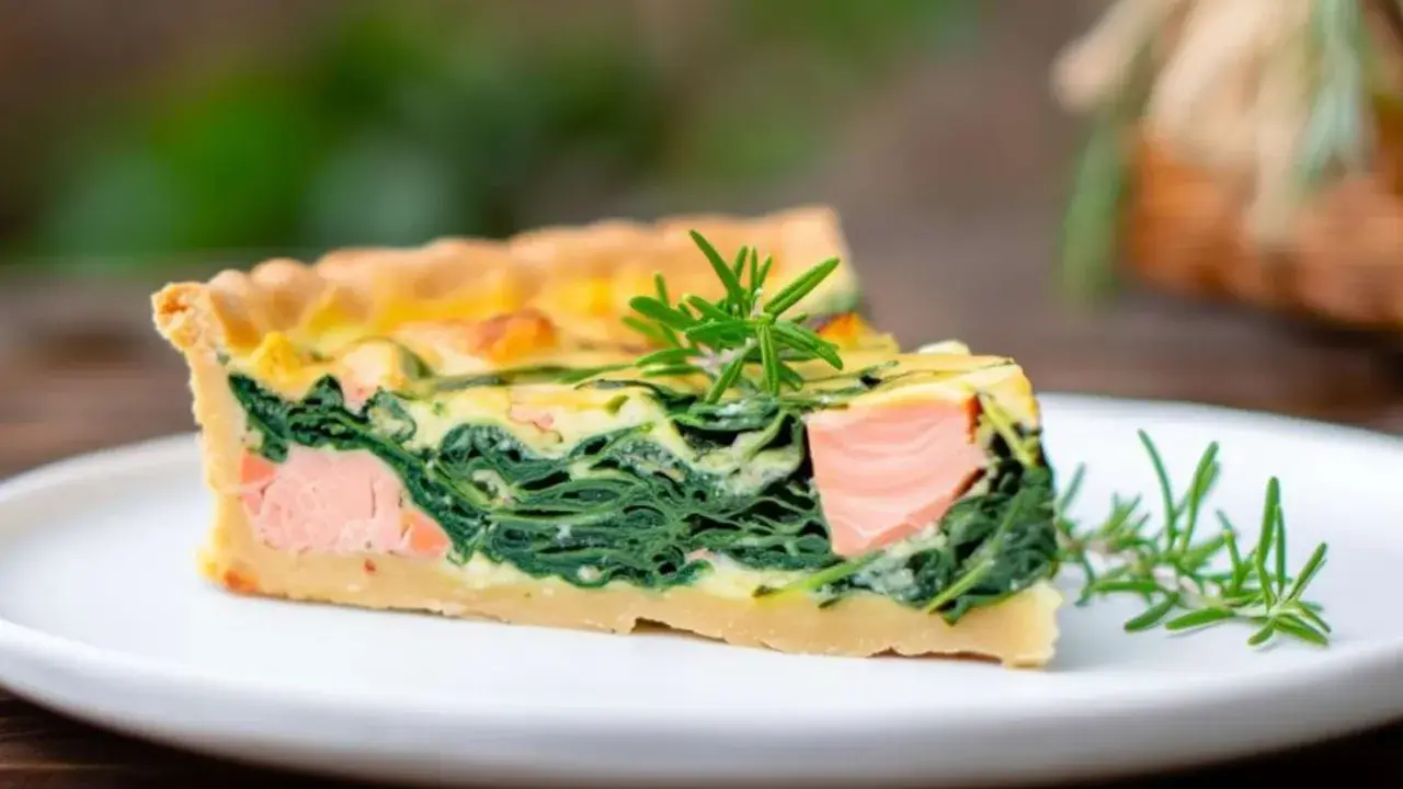 Ein Stück Lachs Spinat Quiche mit Rosmarin garniert auf einem weißen Teller.