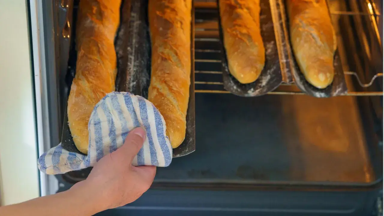 Hand mit Topflappen holt goldbraune Baguettes aus dem Ofen. Der Duft von frisch gebackenen Baguettes liegt in der Luft.
