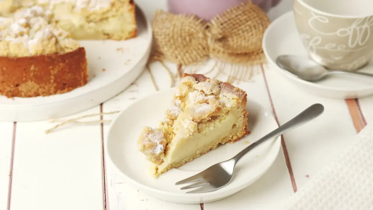 Ein Stück Streuselkuchen mit Pudding auf einem Teller, daneben eine Kaffeetasse und ein ganzer Kuchen.