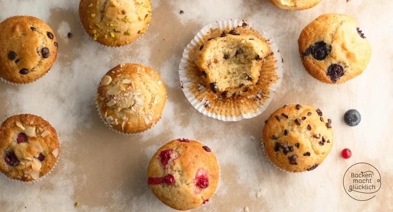 Verschiedene Muffins, darunter Schoko- und Blaubeermuffins. Ein Muffin ist angeknabbert. Die Frage "wie lange muffins backen" wird hier visuell beantwortet: Sie sind perfekt goldbraun.