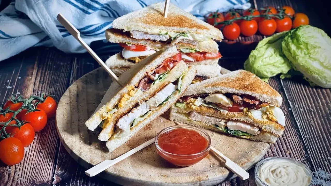 Ein gestapelter Club Sandwich mit Hühnchen, Speck, Ei und Tomaten, serviert mit Ketchup und Mayonnaise.