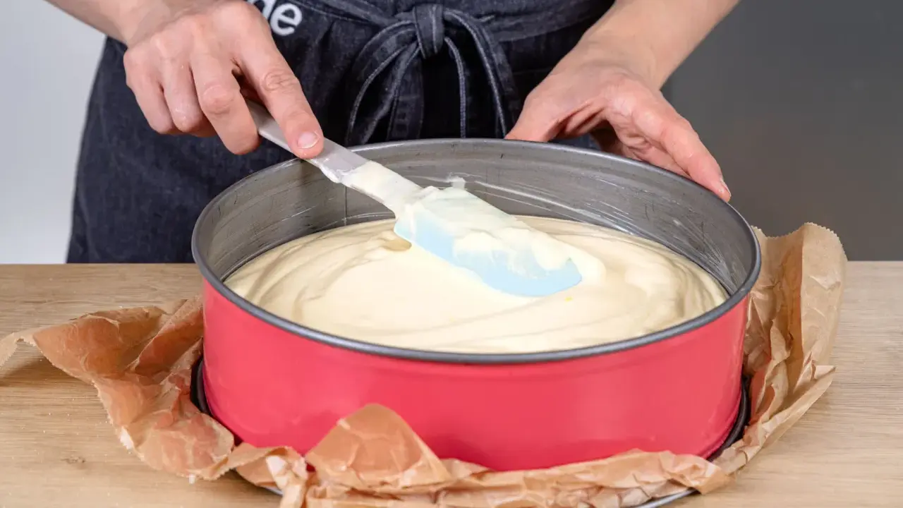 Hände verteilen den Teig für einen Joghurtkuchen mit 500 g Joghurt in einer roten Springform.