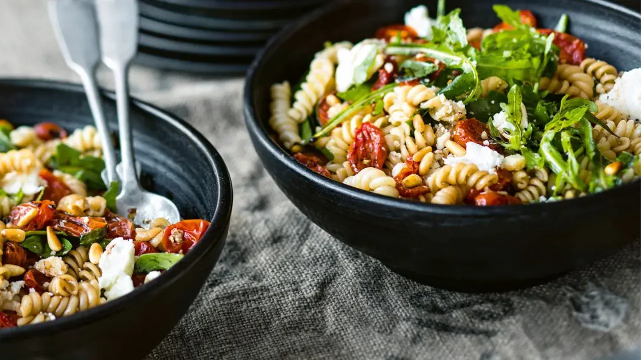 Zwei Schalen mit einem köstlichen nudelsalat mit getrockneten tomaten, garniert mit Rucola und Pinienkernen.