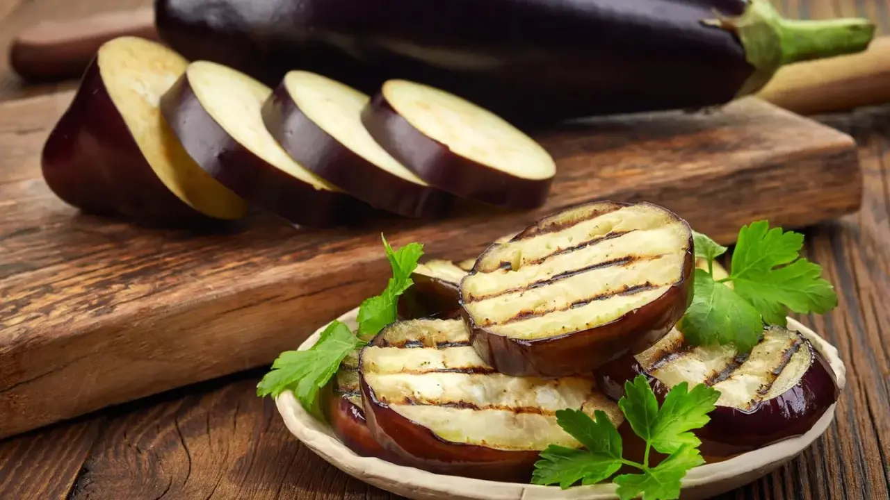 Gegrillte Auberginenscheiben mit Schale, garniert mit Petersilie, liegen auf einem Teller. Daneben liegen weitere Scheiben und eine ganze Aubergine.