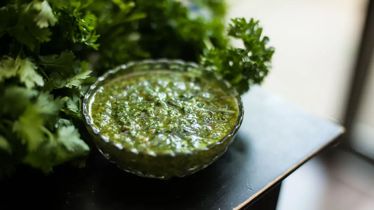 Frische Kräuter umgeben eine Schale mit leuchtend grünem Chimichurri. Was ist Chimichurri? Eine würzige argentinische Sauce aus Petersilie, Knoblauch und Essig.