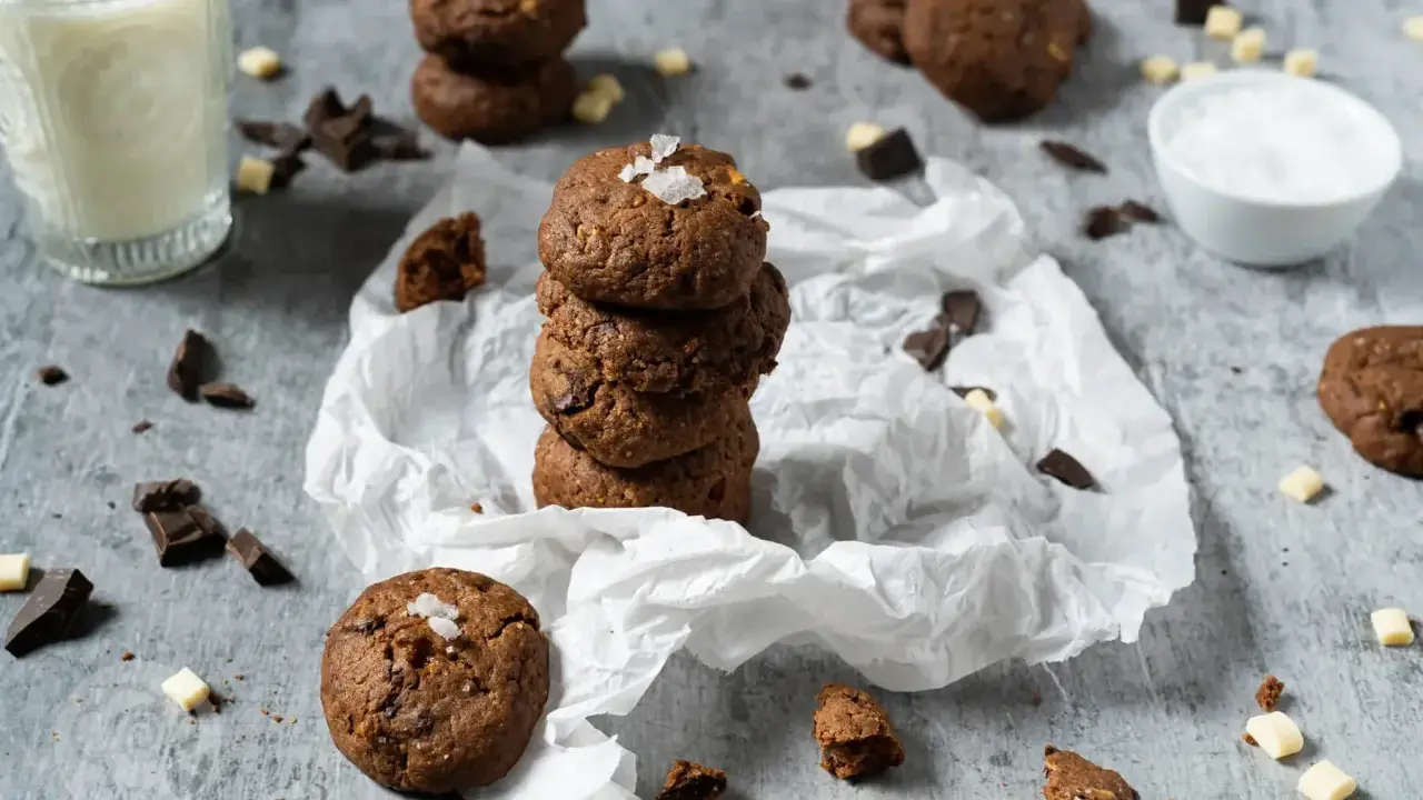 Gestapelte triple chocolate cookies mit Meersalzflocken, umgeben von Schokoladenstückchen und einem Glas Milch.
