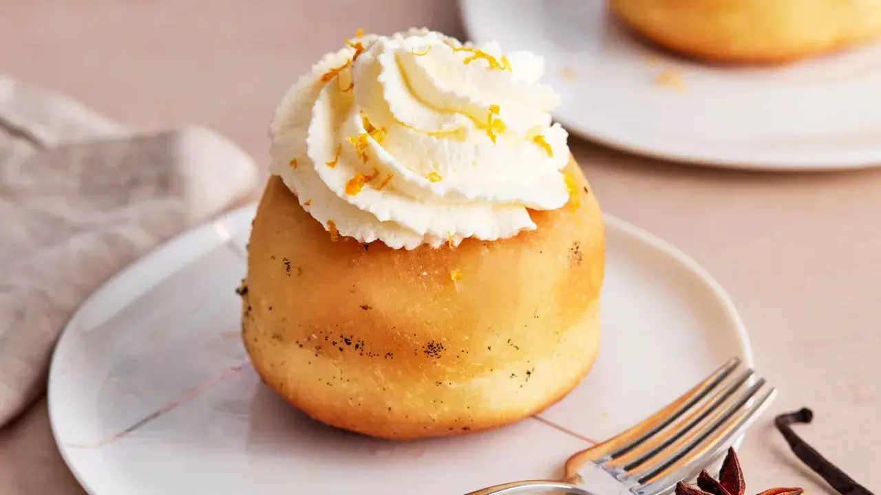 Ein köstlicher Baba au Rhum, gekrönt mit Schlagsahne und Orangenzesten, wartet auf einem Teller. Daneben liegen Sternanis und Vanilleschote.