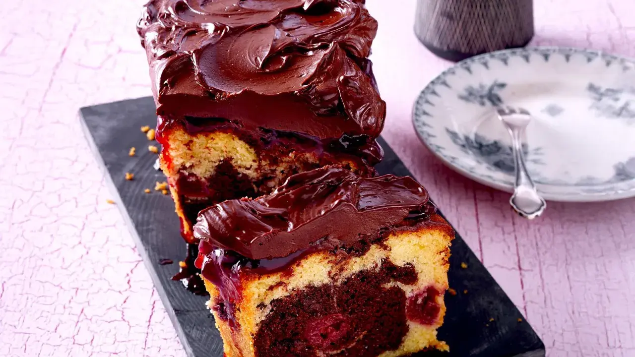 Saftiger Marmorkuchen mit Kirschen, überzogen mit dunkler Schokoladenglasur. Ein Stück ist bereits angeschnitten und zeigt die köstliche Marmorierung.
