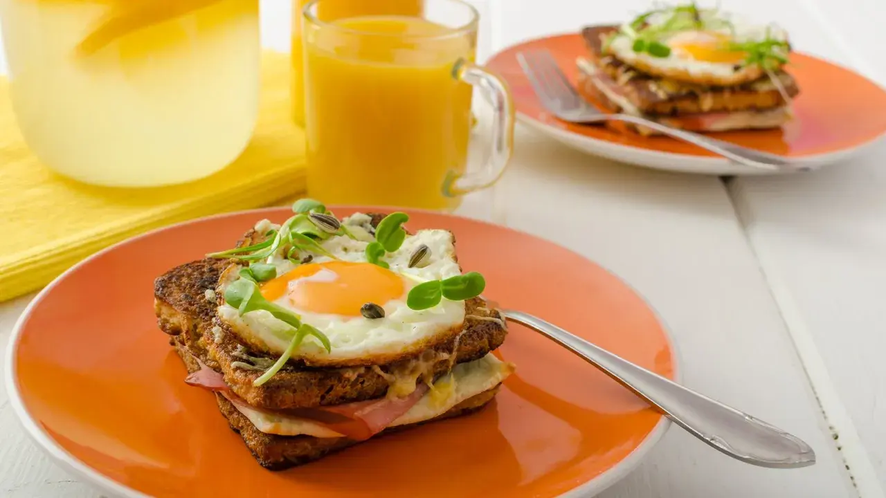 Ein köstlicher Croque Madame mit Spiegelei, Schinken und Käse auf einem orangefarbenen Teller, daneben Saft und Wasser mit Zitrone.