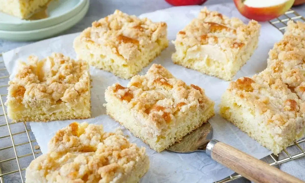 Saftiger Apfelkuchen vom Blech mit knuspriger Streuselhaube. Ein Stück wird gerade mit einem Kuchenheber herausgenommen.