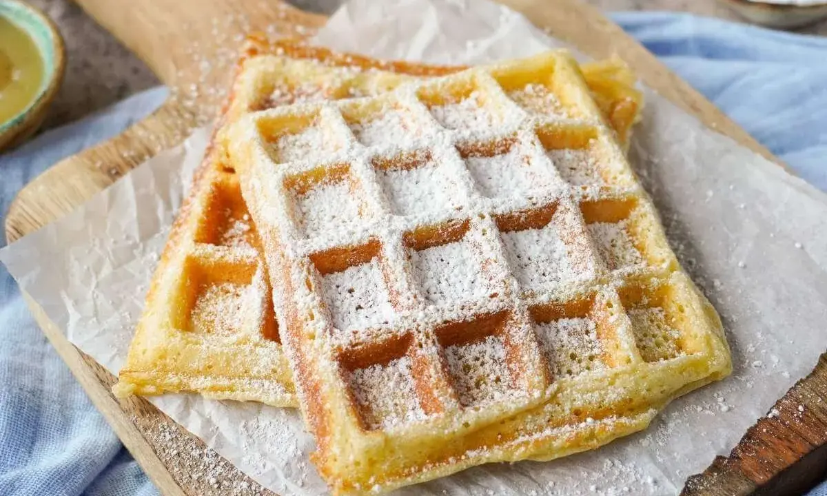 Zwei goldbraune belgische Waffeln, bestäubt mit Puderzucker, liegen auf Backpapier auf einem Holzbrett.