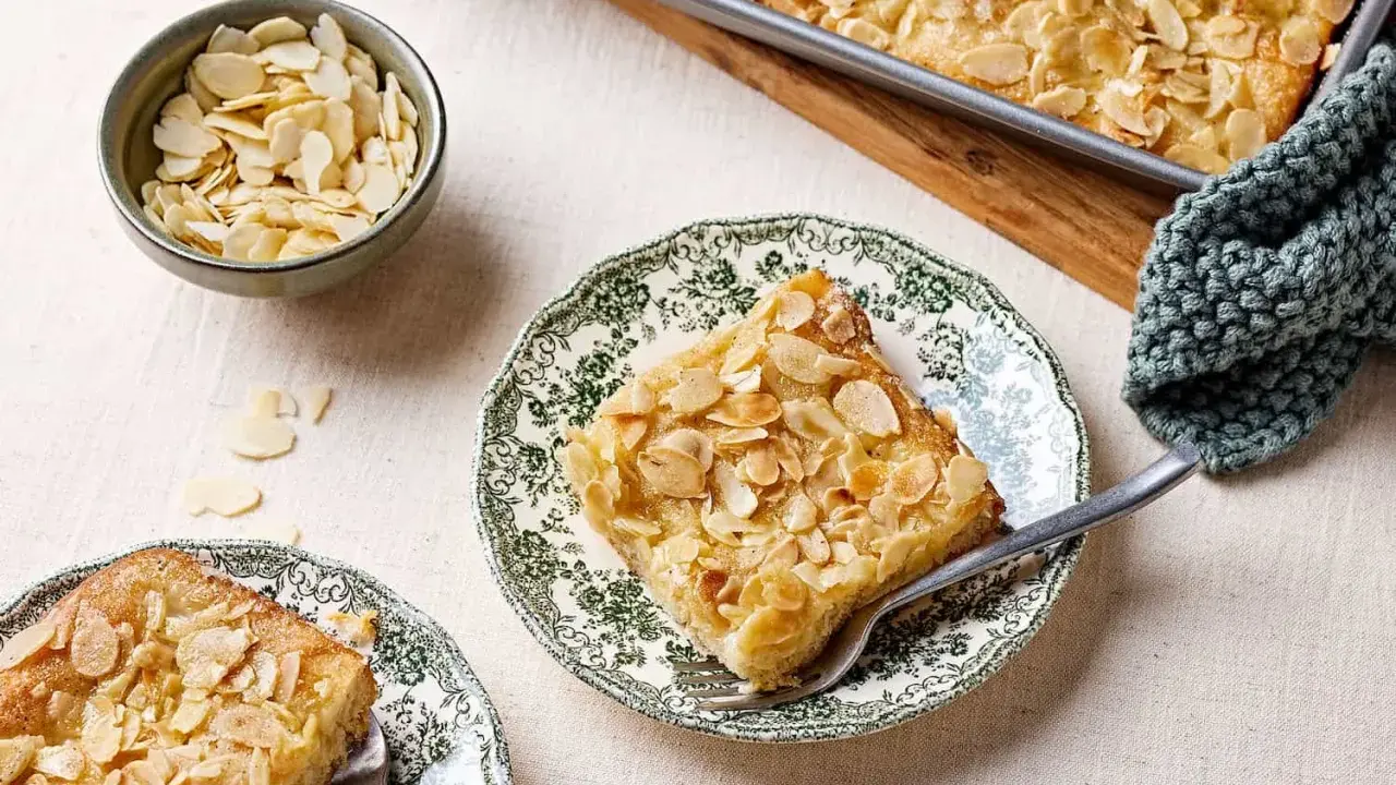 Ein Stück Butterkuchen mit Mandelblättchen auf einem Teller, daneben eine Schüssel mit Mandeln. Perfekt für ein Butterkuchen Rezept.