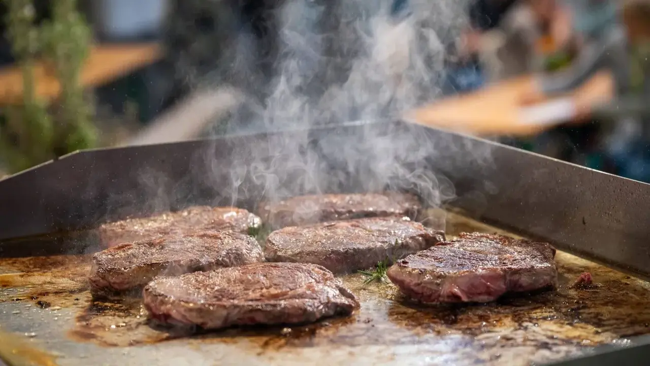 Mehrere saftige Steaks brutzeln auf einer heißen Platte. Dampf steigt auf und zeigt, wie man ein Steak richtig braten kann.
