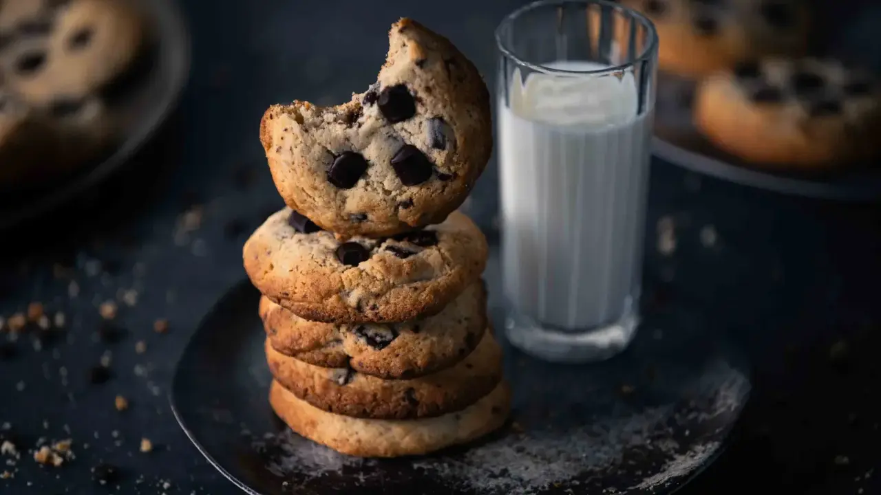 Ein Stapel amerikanischer Cookies mit Schokoladenstückchen, daneben ein Glas Milch. Ein Cookie ist angeknabbert.