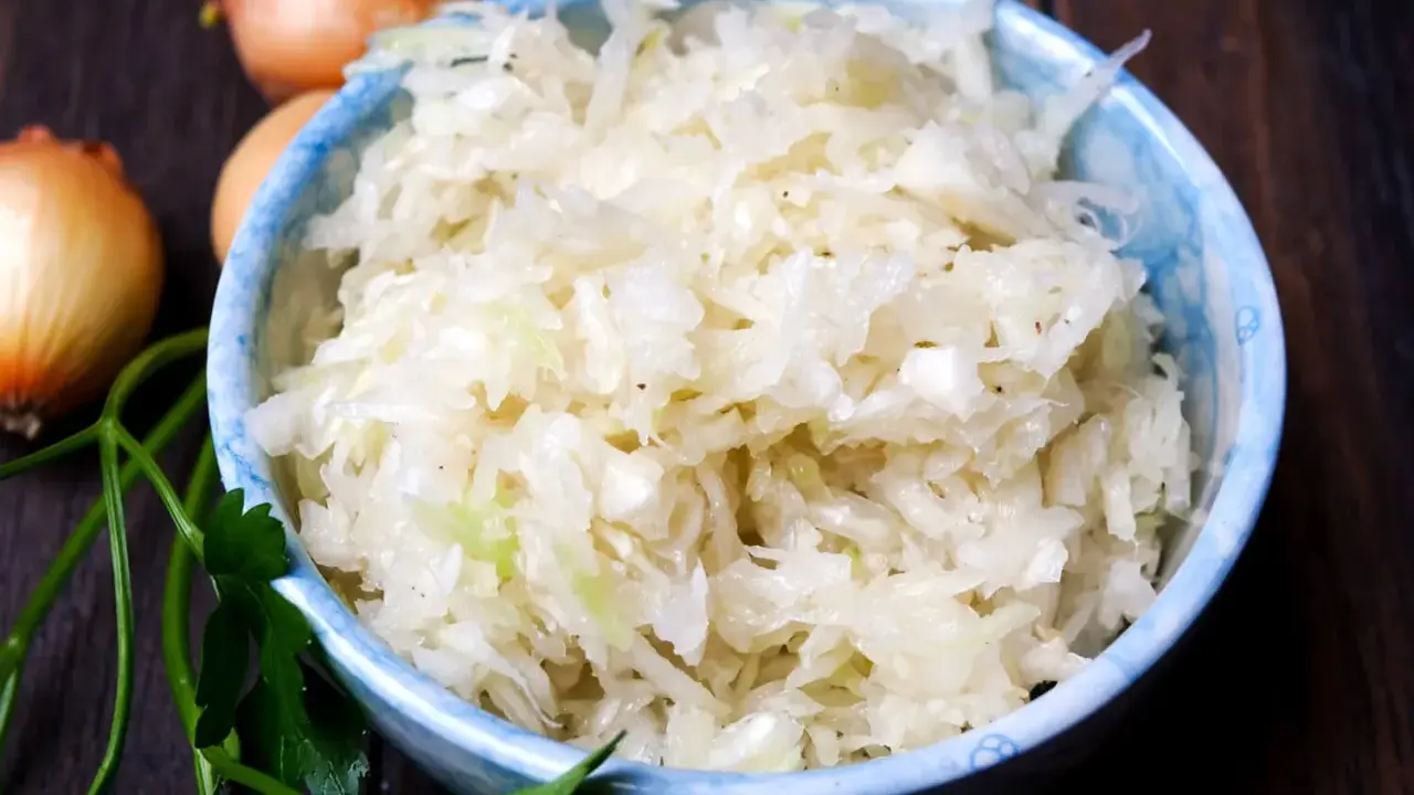 Schüssel mit frisch zubereitetem Krautsalat, daneben Zwiebeln und Petersilie. Perfekt, um Krautsalat selbst zu machen!