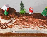 Eine festliche bûche de noël deutsch, verziert mit Weihnachtsmann, Bäumen und einem Haus.