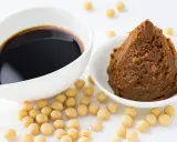 Miso-Paste und Sojasauce in weißen Schalen, umgeben von Sojabohnen. Eine köstliche Alternative zu Hefeflocken für herzhafte Gerichte.