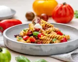 Ein köstlicher italienischer Nudelsalat mit Fusilli, Tomaten, Mozzarella und Basilikum auf einem grauen Teller, umgeben von frischen Tomaten.