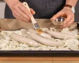 Bratwurst im Backofen: Zwiebelringe auf Blech, darauf vier Würste. Mit Pinsel wird eine Wurst mit Flüssigkeit bestrichen.
