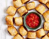 Goldbraune Würstchenschnecken mit Sesam, daneben eine Schale mit roter Chutney-Sauce. Perfekt, um schnelles Fingerfood am Vortag zuzubereiten.