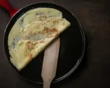 Ein goldbrauner Crêpe wird in einer Pfanne mit rotem Griff gefaltet. Das **Crêpes Teig Rezept Original** gelingt so perfekt.