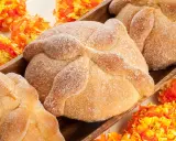 Pan de muerto, süßes Brot mit Zucker bestreut, liegt in einer Holzschale, umgeben von bunten Papierblumen.