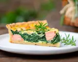 Ein Stück Lachs Spinat Quiche mit Rosmarin garniert auf einem weißen Teller.