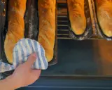 Hand mit Topflappen holt goldbraune Baguettes aus dem Ofen. Der Duft von frisch gebackenen Baguettes liegt in der Luft.