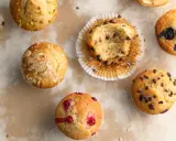 Verschiedene Muffins, darunter Schoko- und Blaubeermuffins. Ein Muffin ist angeknabbert. Die Frage "wie lange muffins backen" wird hier visuell beantwortet: Sie sind perfekt goldbraun.