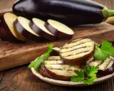 Gegrillte Auberginenscheiben mit Schale, garniert mit Petersilie, liegen auf einem Teller. Daneben liegen weitere Scheiben und eine ganze Aubergine.