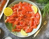 Frisch gebeizter Lachs auf einem Teller, garniert mit Zitronenscheiben und Dill. Daneben eine Schale mit Koriandersamen.