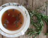 Eine Tasse Kräutertee mit Estragon-Verwendung. Frische Kräuter liegen daneben auf Holz.
