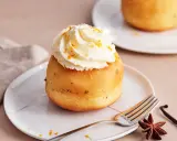 Ein köstlicher Baba au Rhum, gekrönt mit Schlagsahne und Orangenzesten, wartet auf einem Teller. Daneben liegen Sternanis und Vanilleschote.
