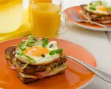 Ein köstlicher Croque Madame mit Spiegelei, Schinken und Käse auf einem orangefarbenen Teller, daneben Saft und Wasser mit Zitrone.