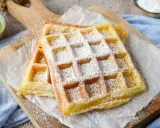 Zwei goldbraune belgische Waffeln, bestäubt mit Puderzucker, liegen auf Backpapier auf einem Holzbrett.