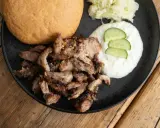Ein Teller mit gegrilltem Fleisch, Tzatziki, Gurkenscheiben, Krautsalat und einem Brötchen – perfekt für ein leckeres Gyros Rezept.