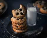 Ein Stapel amerikanischer Cookies mit Schokoladenstückchen, daneben ein Glas Milch. Ein Cookie ist angeknabbert.
