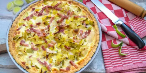 Herzhafte Quiche mit Lauch und Speck, perfekt für herzhafte Quiche Rezepte. Daneben liegen Lauch, ein Messer und ein Nudelholz.