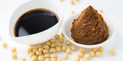 Miso-Paste und Sojasauce in weißen Schalen, umgeben von Sojabohnen. Eine köstliche Alternative zu Hefeflocken für herzhafte Gerichte.