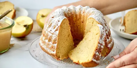Ein saftiger Kuchen mit Apfelmus, bestäubt mit Puderzucker. Daneben liegen Äpfel und ein Glas Apfelmus.