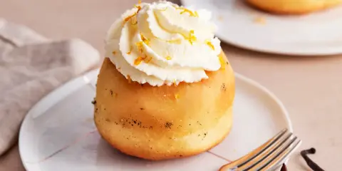 Ein köstlicher Baba au Rhum, gekrönt mit Schlagsahne und Orangenzesten, wartet auf einem Teller. Daneben liegen Sternanis und Vanilleschote.